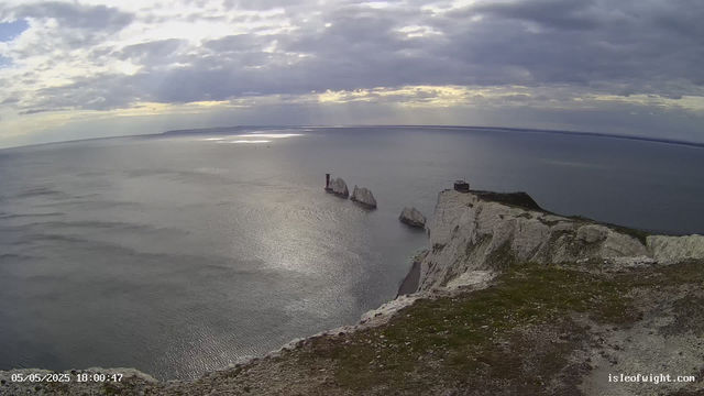 A view from a webcam showing white chalk cliffs along the coastline with a grassy and rocky top edge. In the ocean below, several large rock formations rise out of the water, with one having a tower-like structure on it. The sky is partly cloudy with the sun breaking through the clouds, creating reflections on the water surface. The timestamp on the image reads "05/05/2025 18:00:47" and the website "isleofwight.com" is visible in the bottom right corner.