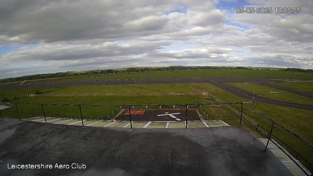A wide view of an airfield on a cloudy day, with a motorcycle parking area in the foreground, a grassy field stretching into the distance, and runways visible on the horizon. There is a small sign and a flag near the parking area, and the sky is partly cloudy with patches of blue. The timestamp in the upper right corner reads "05-05-2025 18:00:55," and the bottom left corner has the text "Leicestershire Aero Club."
