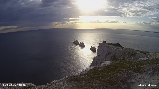 A view from a high vantage point showing white chalk cliffs overlooking the ocean. The sun is setting or rising, casting a golden light on the water. Three large sea stacks stand near the base of the cliffs, and a small building is visible on top of one of the stacks. The sky is partly cloudy, with patches of sunlight breaking through, illuminating the scene. The water appears calm with gentle ripples, and there's a distant sailboat visible on the horizon. The timestamp indicates the photo was taken on May 5, 2025, at 19:00:31, and the URL "isleofwight.com" is displayed in the bottom right corner.