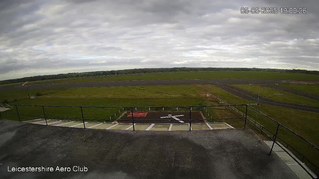An outdoor view of an open grassy area with a runway or taxiway extending into the distance, under a cloudy sky. There are some painted markings on the ground near the foreground, and a small flag on a pole to the left. The image is taken from a balcony or elevated platform with a black railing. The bottom left corner displays the text "Leicestershire Aero Club." The timestamp in the top right corner reads "05-05-2025 19:00:26."