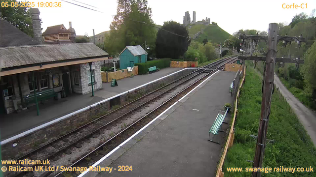 A small train station platform with a historic stone building on the left, featuring a pitched roof and chimney. There are green benches, a sign, and a yellow fence. Railway tracks run along the platform, with a control box or electrical cabinet near the right side. In the background, there is a grassy hill with a castle ruin on top, and a wooded area with trees. The sky is clear, and the overall scene is peaceful with greenery and old structures.