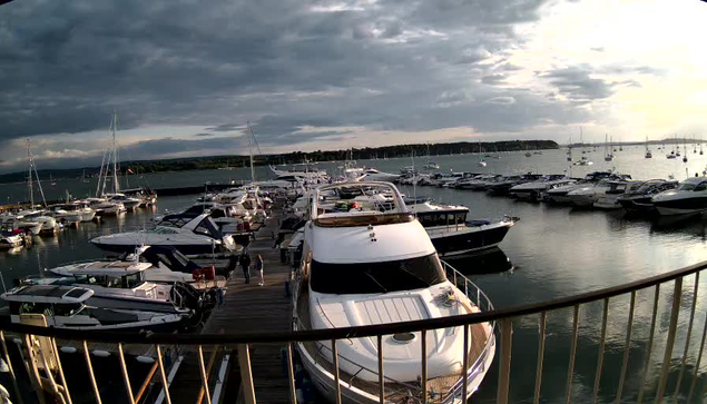 A marina filled with numerous yachts and smaller boats docked along wooden piers. The scene is set during a cloudy day with a mix of dark and light clouds, and there is a faint glow of the sun on the horizon. The water is calm, and a railing runs across the foreground of the image. There are a few people walking on the dock near the boats.