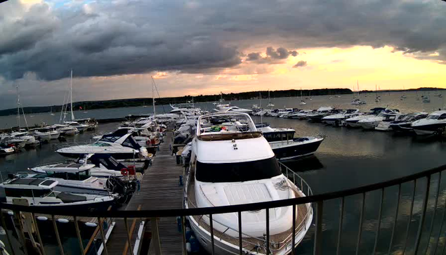 A marina filled with numerous boats and yachts docked along wooden piers under a cloudy sky during sunset, with dark storm clouds on the left and a warm glow on the horizon.