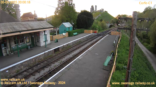 A small railway station platform with benches and a stone building with a corrugated roof. A green shed and a fence with bunting are visible behind the building. There are two railway tracks running parallel, with a white line marking the edge of the platform. An old wooden utility pole with wires stands to the right, and a narrow grassy pathway runs alongside it. In the background, there is a hill with a castle or ruins on top, surrounded by trees and greenery. The overall scene appears to be taken during early evening, with the date and time stamp showing 05-05-2025 at 20:00:18.