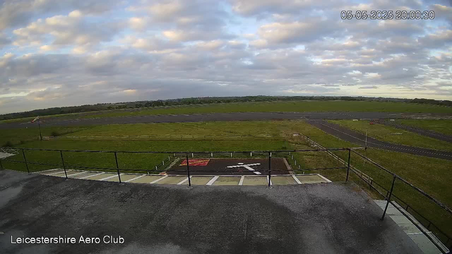 A view from a webcam at Leicestershire Aero Club shows an open grassy area with a small runway or taxiway in the distance. The sky is partly cloudy with patches of blue visible. There's a flagpole on the left side, and the foreground features a fenced area with a helicopter landing pad marked by an 'H' and an arrow. The timestamp in the upper right corner reads "05-05-2025 20:00:30."