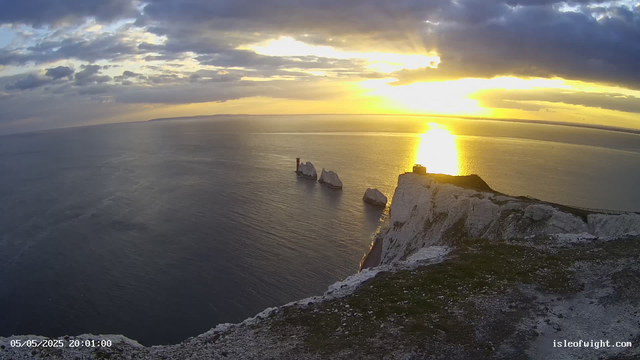 A view from a webcam overlooking the ocean at sunset. White chalk cliffs in the foreground with a small building and a fence on top. The sea is calm, reflecting the golden light of the sunset, with three large sea stacks extending from the water near the cliffs. The sky is partly cloudy, with the sun low on the horizon casting a bright reflection on the water. The timestamp shows "05/05/2025 20:01:00" and the website "isleofwight.com" is visible in the corner.