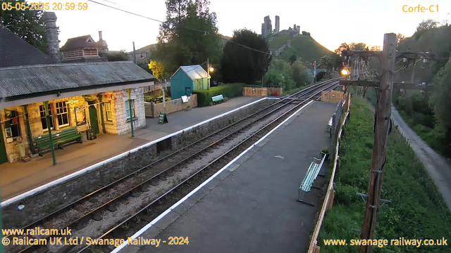 A small railway station platform at sunset, with benches and a small green building. There is a stone station building with green posts and a window, and a sign that says "Way Out." The railway tracks run parallel to the platform, curving slightly to the right in the distance. A wooden utility pole with attached electrical wires is on the right side. In the background, there are trees, a grassy hill with ruins or old structures on top, and a sunset sky. A narrow pathway runs alongside the tracks, bordered by greenery. The timestamp indicates the date and time as May 5th, 2025, at 8:59 PM.