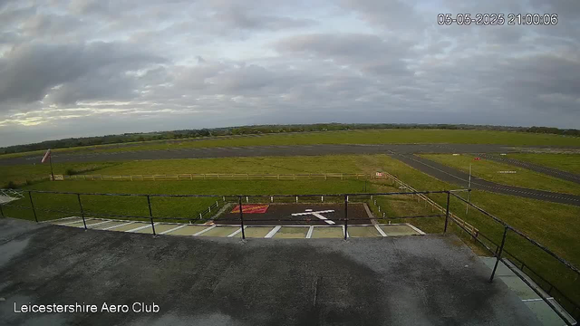 A wide view of an airport runway and grassy fields under a cloudy sky, taken from a high vantage point. In the foreground, there is a small parking area with marked parking spaces and a fenced platform. The date and time stamp shows "05-05-2025 21:00:06," and the location is labeled as "Leicestershire Aero Club."