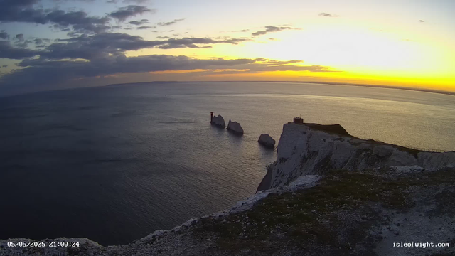 A view of white chalk cliffs overlooking the sea at sunset. There are three sea stacks standing in the water nearby, with the sky partly cloudy and the horizon glowing with yellow and orange hues. The cliffs have patches of grass and a small structure at the top. The scene is calm and picturesque.
