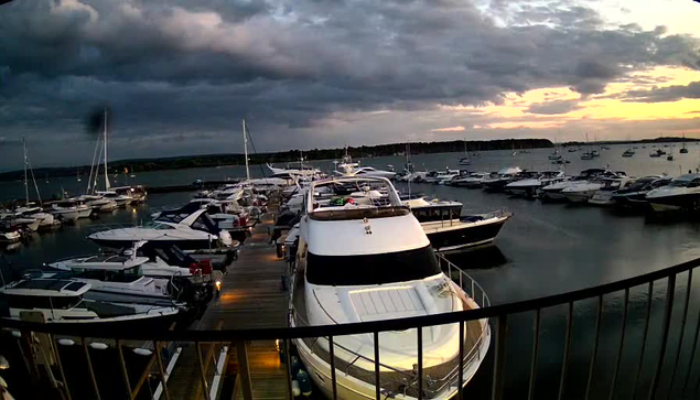 A marina filled with numerous docked boats and yachts during sunset. The sky is partly cloudy with the setting sun casting a warm glow on the horizon, while dark clouds loom overhead. The view is from a balcony with a black railing in the foreground.