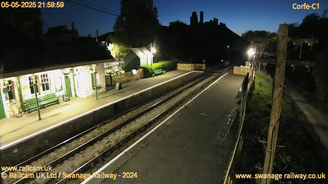A small train station platform at night, illuminated by bright lamps. The platform runs alongside a set of railway tracks, with a small stone building on the left side featuring green doors, windows, and benches. There is a green picnic table outside, with a sign indicating "Way Out." On the right, a wooden fence and poles with electrical wires are visible. The sky is dark, and some trees and building silhouettes are in the background. The timestamp shows "05-05-2025 21:59:59" and the image is from Swanage Railway.