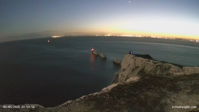 A view of a coastal cliff at dusk or dawn. The sky is darkening or brightening, with a faint glow near the horizon. There are a few illuminated navigation beacons or lights on rocks in the water just below the cliff. The sea is calm, and a distant shoreline with city lights is visible across the water. The timestamp on the image reads "05/05/2025 21:59:56," and the website "isleofwight.com" is noted in the corner.