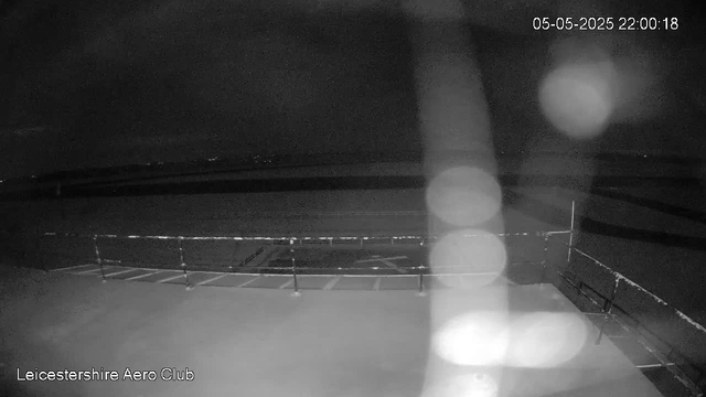 A black-and-white image from a webcam shows an outdoor sports field or track area at night, with barriers or railings in the foreground. The scene is dark, and the image has some blurry light spots or bokeh effects. The top right corner displays the date and time as 05-05-2025 22:00:18. The bottom left corner has the text "Leicestershire Aero Club."