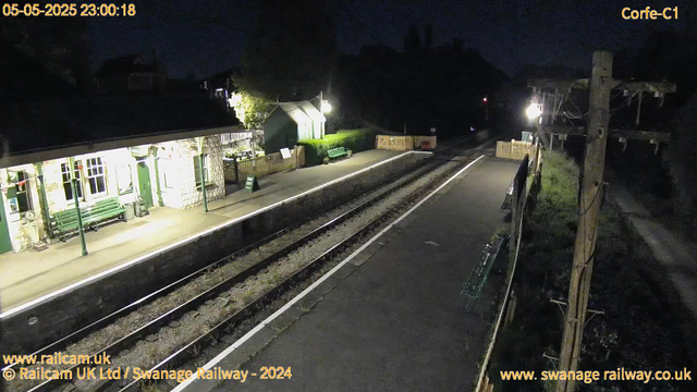 A small train station platform at night with a building on the left, illuminated by a streetlamp. There are a few benches, a sign saying "WAY OUT," and some greenery in the background. To the right, power lines and a tall wooden utility pole are visible. The railway tracks run through the middle of the scene, and the overall atmosphere is quiet and dark.