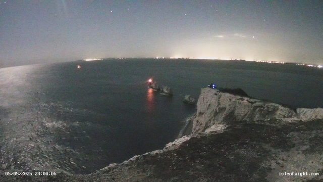 A nighttime view of cliffs and the ocean, with small boats anchored near the shoreline. The horizon features a faint glow of city lights in the distance, and the sky is clear with visible stars. The timestamp on the image reads "05/05/2025 23:00:06," and the website "isleofwight.com" is noted in the bottom right corner.