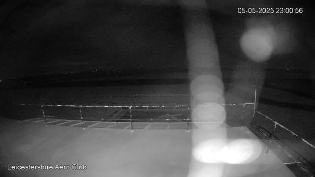 A black-and-white webcam image shows an outdoor area at night, with a view of a fenced-off section that appears to be a runway or open space. The scene is dark with some bright light reflections creating circular bokeh effects. The top right corner displays the timestamp "05-05-2025 23:00:56," and the bottom left corner shows the text "Leicestershire Aero Club."