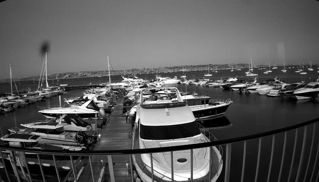 A black-and-white photograph of a marina shows multiple boats and yachts docked along wooden piers. The water is calm, and several boats are moored on both sides of the dock, with larger yachts positioned in the foreground. In the background, there is a distant shoreline with low hills or land formations. The scene is viewed from a vantage point behind a curved railing at the top edge of the image.
