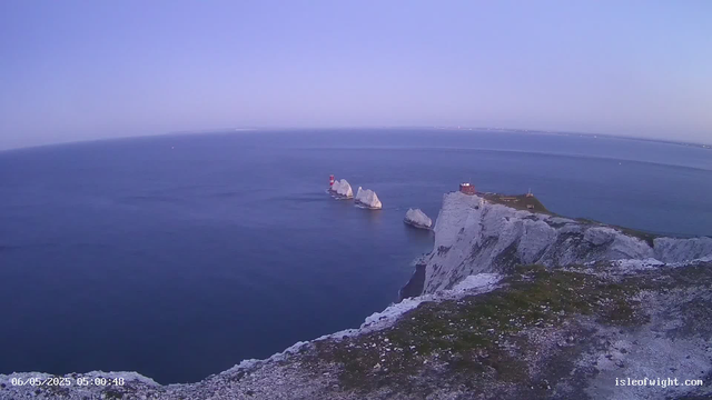 A wide aerial view of white chalk cliffs rising from the dark blue sea under a clear, pale blue sky. Several large rock formations and sea stacks are visible near the shoreline, with a red and white lighthouse on one of the sea stacks. A small building with a red roof is located on top of the largest cliff. The coastline extends in both directions, fading into the horizon. The bottom left corner shows a timestamp, and the bottom right corner displays a website URL.