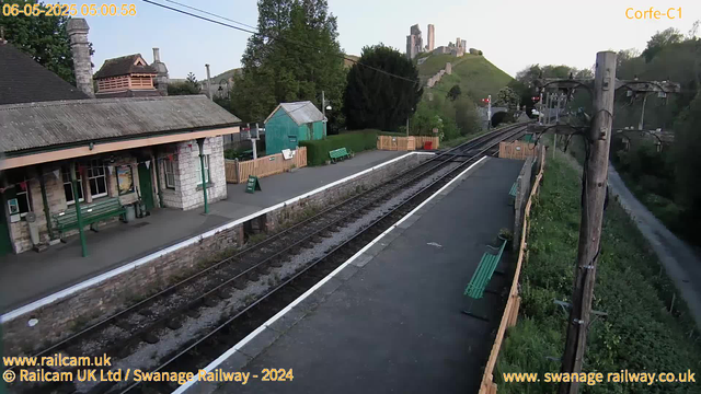 A small railway station platform with a building on the left side, featuring green benches and a trash bin. There are rail tracks running through the center of the image. In the background, there are trees, a small green shed, and a hillside with ruins of an old castle. There are power lines and poles along the platform, and the time and date stamps in the top left corner indicate early morning on June 5th, 2025. The image is captured by a webcam operated by Swanage Railway.