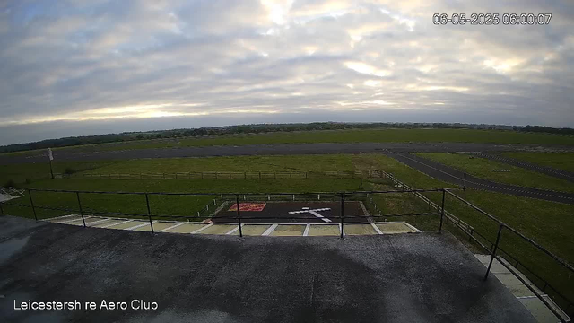 A wide view of an outdoor airfield shows a cloudy sky with patches of sunlight breaking through. In the foreground, there is a platform with a railing, and an area marked with lines and a helicopter landing pad. The surrounding grassy fields stretch toward a taxiway and runways in the distance, with a line of trees on the horizon. The text "Leicestershire Aero Club" is visible at the bottom left corner, and the date and time "06-05-2025 06:00:07" are displayed at the top right corner.