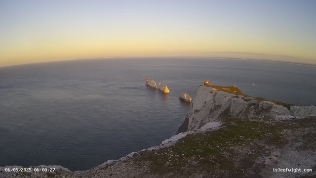 An aerial view of white chalk cliffs with patches of green grass on top, jutting out into the sea during sunrise or sunset. The ocean below is calm with gentle waves, and a sailboat is visible on the water. The sky has a gradient from light yellow near the horizon to pale blue higher up. A small lighthouse or navigation marker stands on one of the rock formations in the sea. The scene is peaceful and illuminated by warm, soft light.
