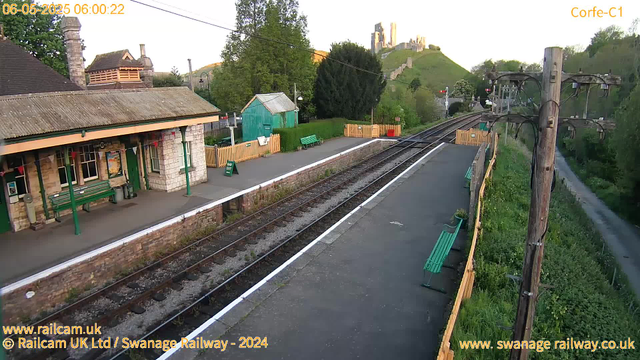 A small train station platform with green benches, a green-painted building, and a stone building with a corrugated roof. There are railway tracks running parallel to the platform, with a row of wooden fencing and a sign that says "Way Out" near the building. Overhead, there are utility poles and wires, and in the background, a green hillside with a castle or ruins on top. The scene is illuminated by early morning sunlight.