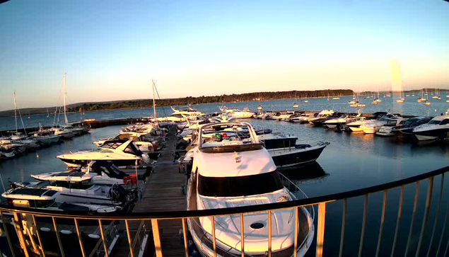 A marina filled with various boats and yachts docked along a wooden pier during sunset. The sky is clear with a gradient of blue and orange hues, and the calm water reflects the boats and surrounding scenery. A curved metal railing is visible in the foreground.