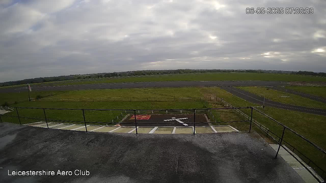 An overcast sky with gray clouds over a large open grassy area and a small airport runway. The view is from a webcam positioned on a building, overlooking the tarmac and surrounding fields. The scene appears calm, with no visible aircraft or people, and the environment looks early in the morning. A timestamp on the image reads "06-05-2025 07:00:09," and text in the lower left corner identifies the location as "Leicestershire Aero Club."