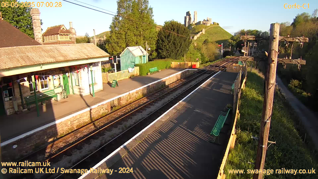 A small train station with a platform, green benches, and a blue shed. There are electrical lines and wooden telegraph poles next to the tracks. In the background, there is a hill with a castle ruin on top, surrounded by green trees and bushes. The scene is bathed in early morning sunlight.