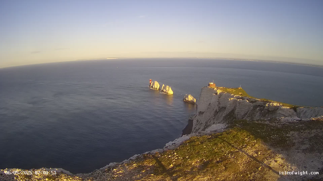 A wide view of white chalk cliffs extending into the ocean, with several large sea stacks offshore. The sun casts a warm light, giving the scene a golden glow. The sky is clear with a soft gradient from light to darker blue, and the calm sea reflects the sky’s colors. The image includes a timestamp in the bottom left corner and a website address in the bottom right corner.