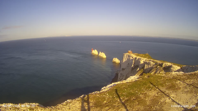 A view of the white chalk cliffs of Étretat in France during sunset, with a lighthouse and a building on top of the cliffs. Several sea stacks are visible in the calm blue waters below, and sailboats are scattered across the horizon in the distance. The sky is clear with a soft gradient from light to darker blue.