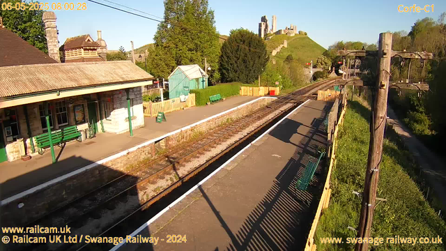 A small train station with a single platform is shown. There is a stone building on the left with a tiled roof and green benches outside. A green fence surrounds a small, turquoise shed in the background, along with some trees and greenery. Railway tracks run parallel to the platform, extending into the distance, with overhead electrical wires and poles alongside. In the background, a hill with a ruin or castle remains is visible atop, with a clear blue sky above. The timestamp indicates the photo was taken on June 5th, 2025 at 8:00 AM.