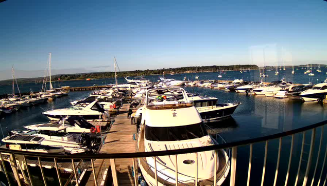 A view of a marina with a wooden dock in the center, filled with various boats and yachts securely moored. The water is calm and reflects the clear blue sky above. In the background, there is a distant shoreline with trees and a few boat masts visible on the horizon. The photo appears to be taken from an elevated position, possibly through a glass window or barrier.