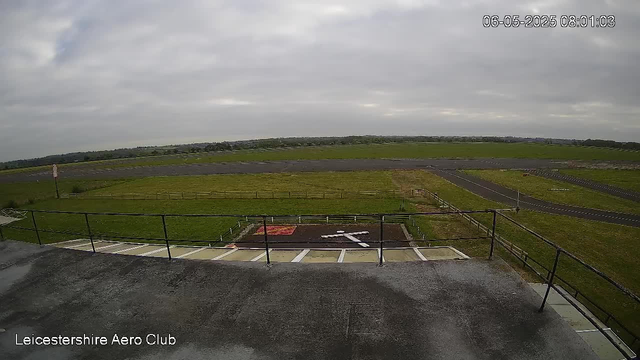 An outdoor view from a webcam showing a cloudy sky over a grassy airfield with taxiways and runways in the distance. In the foreground, there is a small section of a rooftop with a metal railing, and parking spaces are visible below. The image is timestamped "06-05-2025 08:01:03" at the top right corner, and “Leicestershire Aero Club” is written at the bottom left corner.
