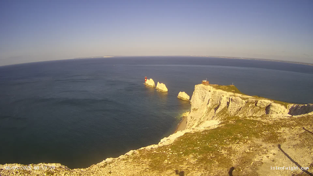 A view from a webcam shows white chalk cliffs extending into the ocean with a lighthouse on the coast. There are rock formations in the water near the cliffs, and the sky is clear with no clouds. The water is calm, with gentle waves, and the scene is brightly lit, suggesting it is daytime. The bottom left corner displays a timestamp, and the bottom right corner has a web address.