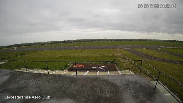 A view from a webcam showing an overcast sky with grey clouds above a grassy airfield. In the foreground, there is a concrete platform with a black metal railing, and underneath the platform are parking spaces. The airfield has runways and taxiways stretching into the distance, with a few small vehicles visible near the edges. The image is labeled "Leicestershire Aero Club" at the bottom left corner and has a timestamp in the upper right corner indicating it was taken on June 5, 2025, at 09:00:04.