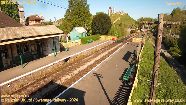 A small train station platform with a stone building on the left that has a tiled roof and a chimney. There are a few green benches along the platform and a sign that says "Way Out." The railway tracks run through the middle of the image, with overhead electric lines supported by wooden poles on the right side. In the background, there are trees, a small green shed, and a hill with ruins or a castle on top. The sky is clear and blue. The image is branded with "railcam.uk" and "Swanage Railway."