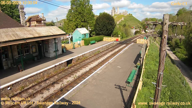 A small train station platform with a building on the left, featuring a tiled roof and two chimneys. The platform has green benches, a small green shed, and some yellow fencing at the far end. Railroad tracks run through the center of the image, with electric poles and wires along the right side. In the background, there is a lush green hillside with a castle ruin perched on top, under a partly cloudy sky with some patches of blue.