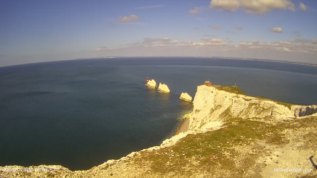 A panoramic view of white chalk cliffs reaching into the sea, with a lighthouse and small building perched on the edge of the cliffs. In the background, the ocean stretches out to the horizon under a partly cloudy sky. There are notable sea stacks close to the cliffs, and the water appears calm. The image captures a bright, sunny day with clear visibility.