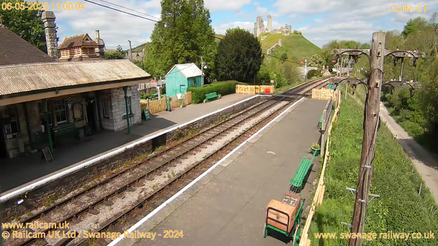 A small vintage train station platform with a building on the left, green benches, and a small turquoise shed. Railroad tracks run through the center, with a grassy hill and castle ruins visible in the background. There are power lines and wooden posts with electric wires on the right side. The sky is partly cloudy with patches of blue. The photo is timestamped May 6, 2025, at 11:00 am.