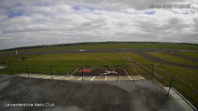 An outdoor scene at Leicestershire Aero Club shows a cloudy sky with scattered clouds. In the foreground, there is a concrete viewing platform with black metal railings and a small parking area with marked spaces. A small white aircraft is parked on the grass near the runway in the middle distance. The surrounding landscape is open with green fields and a few distant trees, with a winding runway running horizontally across the image. The timestamp in the top right corner indicates the date and time as June 5, 2025, at 11:00:22.