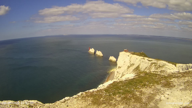 A panoramic view of white chalk cliffs along the coastline, with a small lighthouse and a building on top of the cliffs. In the ocean below, there are large white rock formations, one of which has a small red and white lighthouse. The sky is partly cloudy with patches of blue.