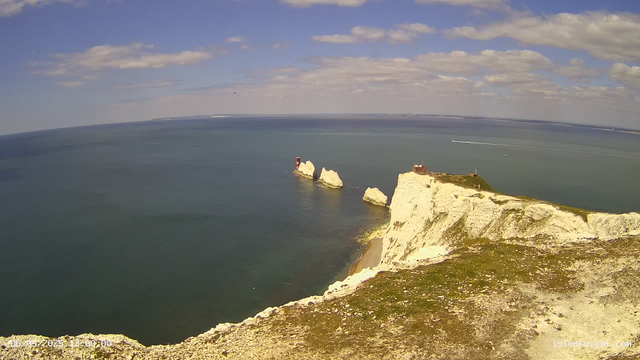 A panoramic view of white chalk cliffs along the coastline extending into the sea, with a lighthouse on the highest point of the cliffs. Several large chalk stacks rise from the water offshore, and a small building is situated at the edge of the cliffs. The sea is calm with a few boats creating gentle wake patterns, and the sky above is partly cloudy with patches of blue.