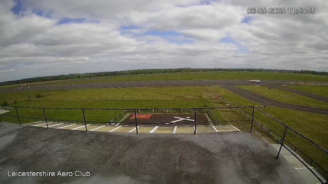 A view from a webcam showing an open grassy area with a small runway or taxiway. There is a grassy field extending into the distance under a cloudy sky. In the foreground, there is a concrete platform with a black metal railing and a few marked parking spaces. The bottom left corner shows the text "Leicestershire Aero Club." The top right corner displays the date and time: 06-05-2025 11:59:55. A small airplane is visible on the runway in the background.