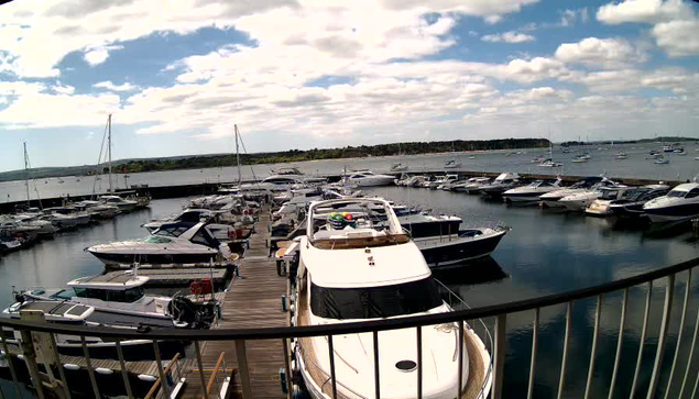A marina filled with numerous boats docked along wooden piers. The water is calm and reflects the boats and the sky. The sky is partly cloudy with patches of blue and white clouds. A curved metal railing is in the foreground, overlooking the docked boats.