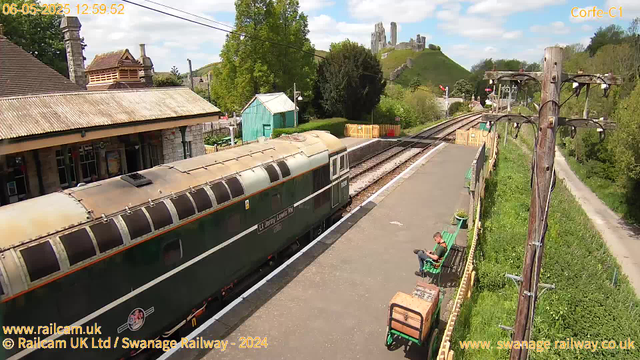 A vintage train with a green body and cream-colored roof is stopped at a small rural station. There are two green benches on the platform, one of which a man is sitting on, using his phone. The platform has a yellow railing, and a wooden utility pole with wires runs alongside it. In the background, there are trees, a small teal-colored building, and a hill with ancient stone ruins on top. The sky is partly cloudy with bright sunlight. A dirt path runs parallel to the railway track on the right side of the image. The timestamp in the top left corner reads "06-05-2025 12:59:52."