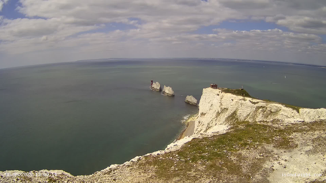 A panoramic view of white chalk cliffs rising above the ocean, with large rock formations near the shore, some topped with structures. The sky is partly cloudy with patches of blue, and sailboats are visible on the water in the distance.