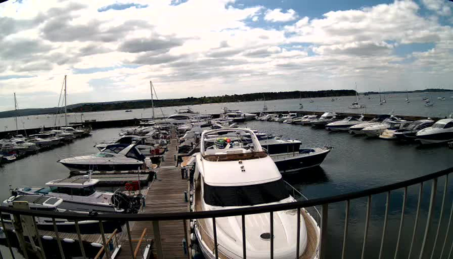 A view from a webcam showing a marina with numerous boats docked along wooden piers. The boats vary in size and style, with some having covered cabins and others open. The water is calm, and the sky above is partly cloudy with patches of blue. In the background, there is a distant shoreline with trees. The image is taken from behind a curved railing in the foreground.