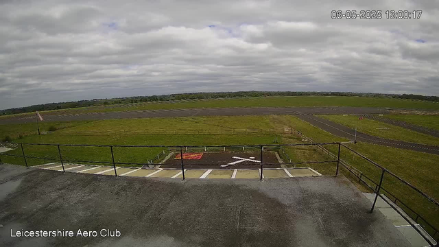 A wide view of an open grassy field under a cloudy sky, taken from a webcam at Leicester Shire Aero Club. The image shows a section of a runway or taxiway in the distance, with a few small structures or signs. The timestamp in the upper right corner indicates the date and time as June 5, 2025, at 1:00 PM. The foreground features a concrete platform with a safety railing and parking spaces below.