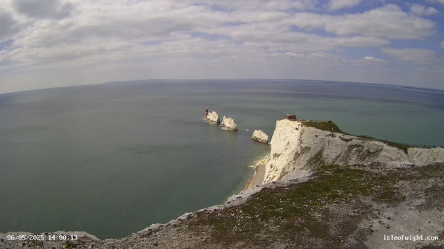 A view from a webcam showing white chalk cliffs along the coastline with grassy patches on top. In the distance, there are two white rock formations rising from the sea, topped with a small red and white lighthouse. The sky is partly cloudy with patches of blue. The water is calm with a greenish hue, and a small part of the rocky shoreline is visible at the bottom of the image. There is a timestamp at the bottom left corner indicating the date and time as 06/05/2025 14:00:13, and a website URL "isleofwight.com" at the bottom right corner.